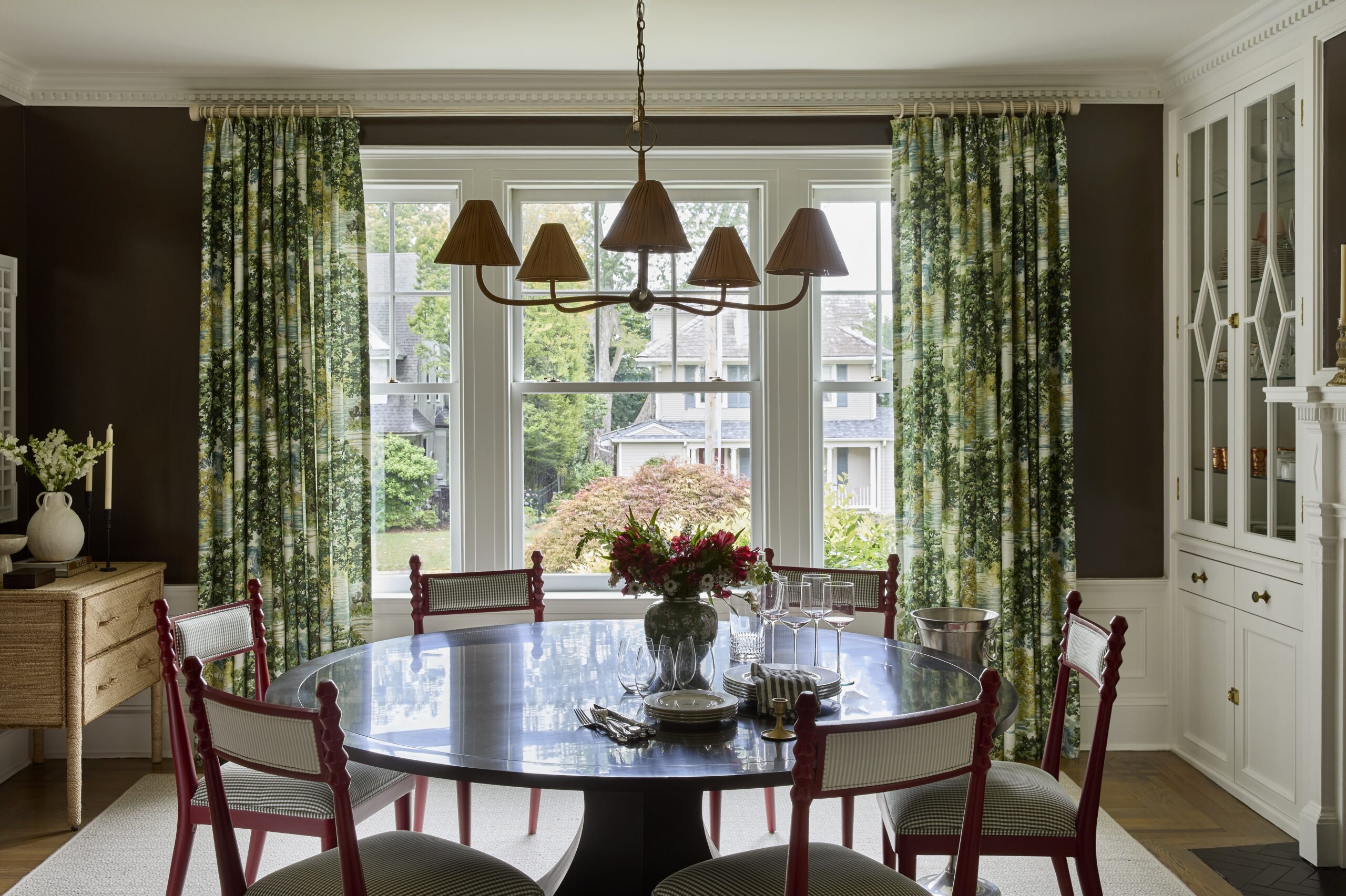 Chocolate brown dining room with red upholstered chairs, white millwork and patterned floral drapery in a Chatham, New Jersey Colonial home.